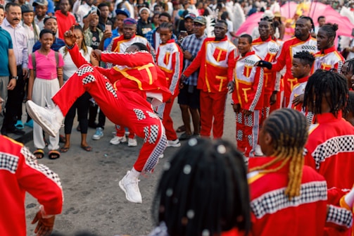A person in vibrant red and checkered attire performs a dynamic leap in front of an audience. The crowd forms a supportive circle, dressed in matching costumes, suggesting a coordinated group performance. Spectators watch attentively, showcasing a lively and festive atmosphere.
