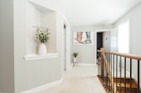 A bright and modern interior hallway with light grey walls and white trim. On the left is an alcove with a glass shelf holding a white vase filled with green foliage. In the center background, there is a colorful abstract painting and below it a small green plant on a thin-legged stand. The right side features a wooden stair railing with black balusters leading downstairs, and there are two doors, one slightly open, revealing a glimpse of another room.