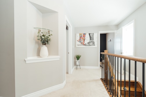 A bright and modern interior hallway with light grey walls and white trim. On the left is an alcove with a glass shelf holding a white vase filled with green foliage. In the center background, there is a colorful abstract painting and below it a small green plant on a thin-legged stand. The right side features a wooden stair railing with black balusters leading downstairs, and there are two doors, one slightly open, revealing a glimpse of another room.
