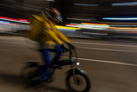 A cyclist wearing a yellow jacket and helmet is riding a bicycle along a city street. The motion blur effect emphasizes speed and movement. Neon lights from nearby buildings create colorful streaks in the background, adding to the sense of urban nightlife.