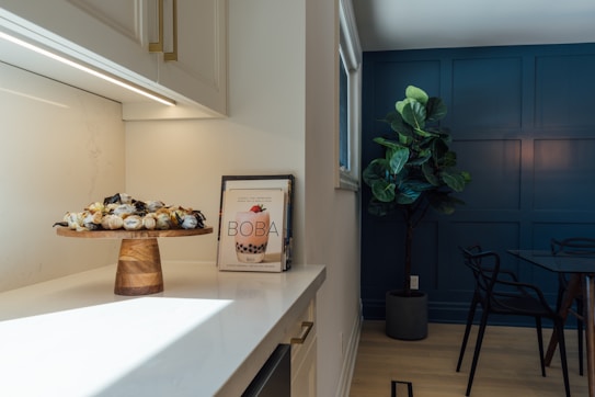 A modern kitchen corner featuring a wooden cake stand with assorted treats, and a cookbook titled 'Boba' on a marble countertop. A large potted plant stands near a dark blue accent wall, adjacent to a dark glass dining table with black chairs.