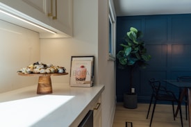 A modern kitchen corner featuring a wooden cake stand with assorted treats, and a cookbook titled 'Boba' on a marble countertop. A large potted plant stands near a dark blue accent wall, adjacent to a dark glass dining table with black chairs.