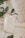 A small beige pouch with a drawstring is placed on a soft, textured fabric. A tag attached to the pouch reads '2024' in bold black numbers. Green leaves from plants frame the pouch, adding a natural element to the composition.