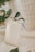A small beige pouch with a drawstring is placed on a soft, textured fabric. A tag attached to the pouch reads '2024' in bold black numbers. Green leaves from plants frame the pouch, adding a natural element to the composition.