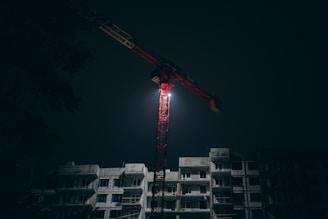 A tall crane illuminated against a dark night sky, positioned above a multi-story construction site. The building is partially constructed and shadowy.