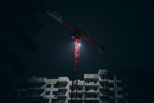 Nighttime shot of a crane with bright lights lifting materials at a construction site.