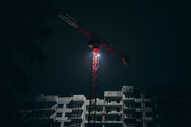 A tall crane illuminated against a dark night sky, positioned above a multi-story construction site. The building is partially constructed and shadowy.