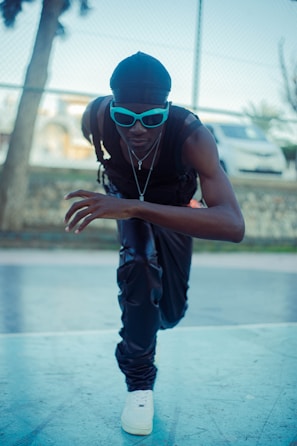 A person in a dynamic, athletic stance appears to be ready to sprint. They are wearing casual athletic clothing and blue-tinted sunglasses. The background suggests an outdoor sports area with a fence, a tree, and a blurred vehicle.