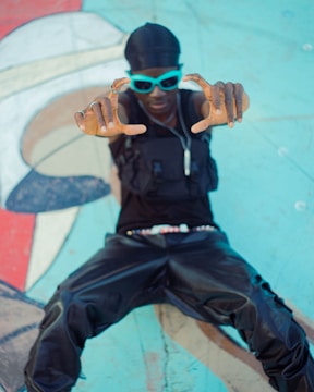 A person wearing blue sunglasses and a black outfit makes a rectangular frame with their fingers against a colorful mural background. They have a stylish and streetwear-inspired look.