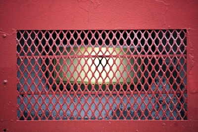 A metal grate with a diamond pattern is positioned over a rectangular structure that houses a light. The grate is painted red, contrasting with the illuminated area behind it, which is a warm yellow-green. The background is a mix of dark and weathered surfaces.