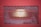 A metal grate with a diamond pattern is positioned over a rectangular structure that houses a light. The grate is painted red, contrasting with the illuminated area behind it, which is a warm yellow-green. The background is a mix of dark and weathered surfaces.