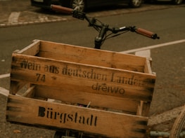 A wooden crate is attached to the handlebars of a bicycle, with German text printed on it. The bicycle is parked on a street, with a car and pavement visible in the background.