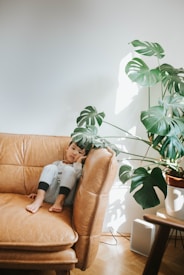 A young child with short hair sits on a tan leather sofa, resting their head on one hand. They are wearing light-colored pajamas or comfortable clothing. Beside the sofa, a large potted Monstera plant with broad leaves casts shadows on the wall. Natural light fills the room, creating a warm and cozy atmosphere.