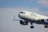 A passenger airplane with jetBlue branding is flying against a partly cloudy sky. The aircraft is in mid-air and its landing gear is visible.