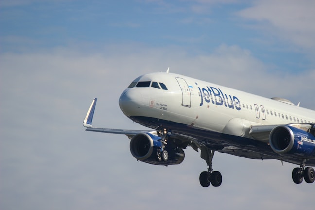 A passenger airplane with jetBlue branding is flying against a partly cloudy sky. The aircraft is in mid-air and its landing gear is visible.