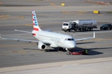 A commercial airplane is being towed on an airport tarmac. The aircraft has American Eagle branding on its fuselage and vertical stabilizer. Nearby, a fuel truck labeled Swissport is stationed, as well as a smaller utility vehicle.