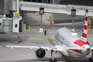 A commercial airplane is parked at a gate, with one airport worker in a vest standing near the front of the airplane. The gate is labeled with the number 12, and there are digital signs, yellow bollards, and other airport ground equipment visible.