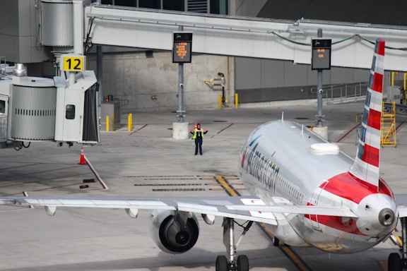 A commercial airplane is parked at a gate, with one airport worker in a vest standing near the front of the airplane. The gate is labeled with the number 12, and there are digital signs, yellow bollards, and other airport ground equipment visible.