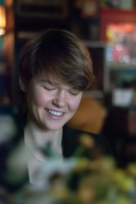 A person with short hair and a gentle smile is captured indoors. The background is softly blurred, featuring warm and muted colors with hints of soft-focus lights, suggesting a cozy or intimate setting. The lighting on the person's face is soft and natural.