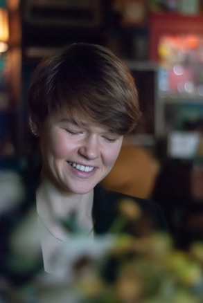 A person with short hair and a gentle smile is captured indoors. The background is softly blurred, featuring warm and muted colors with hints of soft-focus lights, suggesting a cozy or intimate setting. The lighting on the person's face is soft and natural.