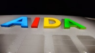 Multicolored letters spelling AIDA are illuminated against a dark background. The letters are vibrant, with blue, red, yellow, and green colors. Below the letters is a section of white material with perforated panels, likely part of a ship or a similar structure.