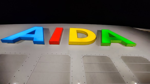 Multicolored letters spelling AIDA are illuminated against a dark background. The letters are vibrant, with blue, red, yellow, and green colors. Below the letters is a section of white material with perforated panels, likely part of a ship or a similar structure.