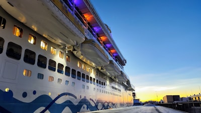 A large cruise ship is docked, featuring multiple levels with illuminated windows and lifeboats situated above. The ship is painted with wavy blue patterns along its white hull. It is evening or early morning, as the sky displays a gradient of blue transitioning into orange and yellow at the horizon.