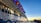A large cruise ship is docked, featuring multiple levels with illuminated windows and lifeboats situated above. The ship is painted with wavy blue patterns along its white hull. It is evening or early morning, as the sky displays a gradient of blue transitioning into orange and yellow at the horizon.