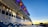 A large cruise ship is docked, featuring multiple levels with illuminated windows and lifeboats situated above. The ship is painted with wavy blue patterns along its white hull. It is evening or early morning, as the sky displays a gradient of blue transitioning into orange and yellow at the horizon.