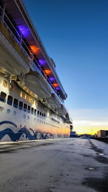 A large cruise ship is docked at a port, with a clear blue sky in the background. The side of the ship is adorned with a stylized wave design and illuminated by colorful lights, including purple and orange hues. Lifeboats are visible, and the reflection of a sunset is seen on the water. The dock area appears empty, providing a serene atmosphere.