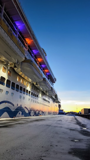 A large cruise ship is docked at a port, with a clear blue sky in the background. The side of the ship is adorned with a stylized wave design and illuminated by colorful lights, including purple and orange hues. Lifeboats are visible, and the reflection of a sunset is seen on the water. The dock area appears empty, providing a serene atmosphere.