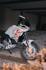 A white motorcycle with branded decals is parked on a gravel surface. It features an intricate design and appears to be well-maintained. The setting is a partially enclosed area with a concrete wall in the background and some dried leaves in the foreground.