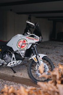 A white motorcycle with branded decals is parked on a gravel surface. It features an intricate design and appears to be well-maintained. The setting is a partially enclosed area with a concrete wall in the background and some dried leaves in the foreground.