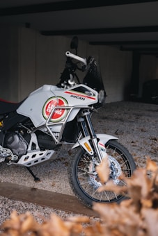 A white motorcycle with branded decals is parked on a gravel surface. It features an intricate design and appears to be well-maintained. The setting is a partially enclosed area with a concrete wall in the background and some dried leaves in the foreground.