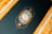 An intricately designed golden frame surrounds a dark background featuring an ornate circular emblem. The emblem includes delicate floral patterns and a central design resembling a compass or clock, crafted with elaborate gold detailing and soft pastel colors.