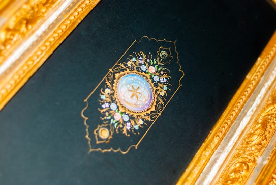 An intricately designed golden frame surrounds a dark background featuring an ornate circular emblem. The emblem includes delicate floral patterns and a central design resembling a compass or clock, crafted with elaborate gold detailing and soft pastel colors.
