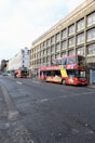 A vibrant red double-decker tour bus and a smaller red bus are parked along a street lined with multi-story buildings. The buses display colorful advertisements with sightseeing themes. Bicycles are parked near the sidewalk, and a motorcyclist is visible further down the road. The scene appears to be in an urban environment with a blend of modern and classic architecture.