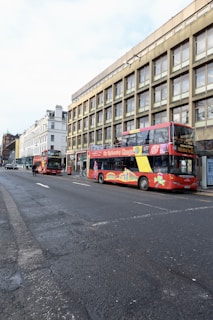 A vibrant red double-decker tour bus and a smaller red bus are parked along a street lined with multi-story buildings. The buses display colorful advertisements with sightseeing themes. Bicycles are parked near the sidewalk, and a motorcyclist is visible further down the road. The scene appears to be in an urban environment with a blend of modern and classic architecture.