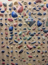 A variety of colorful rock climbing holds are scattered across a vertical climbing wall. The holds are different shapes and sizes, including round, angular, and cylindrical, arranged to create multiple potential climbing routes.