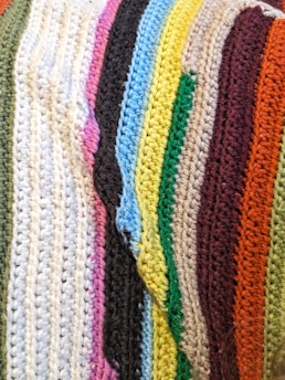 A close-up view of a crocheted fabric featuring multiple colorful stripes aligned next to each other. Each stripe is a different color, showcasing a variety of textures and thicknesses typical of crochet work.