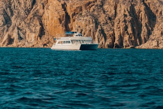 A boat is floating on a deep blue sea with rocky cliffs rising in the background. The cliffs are rugged and have a warm, earthy color. The boat appears to be a small passenger or tour vessel, painted mostly in white.