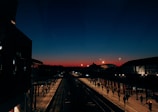 A train station platform is bustling with people walking as the sky transitions from a deep blue to a warm orange glow, indicating either dawn or dusk. The station architecture includes large, illuminated curved roofs and a network of tracks leading into the distance. Silhouettes of passengers are visible, adding to the dynamic scene.
