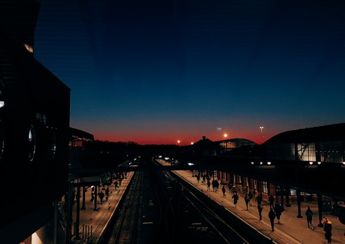A train station platform is bustling with people walking as the sky transitions from a deep blue to a warm orange glow, indicating either dawn or dusk. The station architecture includes large, illuminated curved roofs and a network of tracks leading into the distance. Silhouettes of passengers are visible, adding to the dynamic scene.