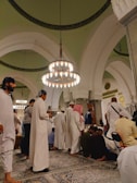 A group of people dressed in traditional attire are gathered in a large, ornate hall featuring high arched ceilings and hanging chandeliers. The setting appears to be a religious or cultural gathering, as people are either standing, sitting, or bowing on intricately patterned rugs.