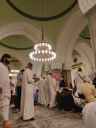 A group of people dressed in traditional attire are gathered in a large, ornate hall featuring high arched ceilings and hanging chandeliers. The setting appears to be a religious or cultural gathering, as people are either standing, sitting, or bowing on intricately patterned rugs.