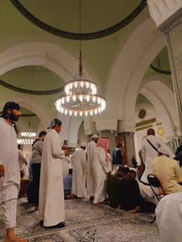 A group of people dressed in traditional attire are gathered in a large, ornate hall featuring high arched ceilings and hanging chandeliers. The setting appears to be a religious or cultural gathering, as people are either standing, sitting, or bowing on intricately patterned rugs.