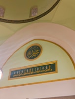 The image portrays an interior architectural detail featuring decorative Islamic calligraphy. The calligraphy is elegantly inscribed on green panels, bordered with gold, set against a light green and beige background. Domes with arched windows are partially visible, and the structure suggests it could be part of a mosque or an Islamic cultural center.