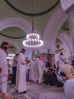 A group of people, dressed in traditional attire, are gathered inside a mosque. The architecture includes large, ornate chandeliers and intricately designed arches. Some individuals are standing, while others are praying on a patterned carpet. The interior walls and ceiling feature decorative calligraphy.