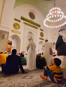 A gathering of people inside a mosque, with intricate Islamic architecture featuring Arabic calligraphy and a large chandelier hanging from the ceiling. Many of the attendees are sitting on a patterned carpet. Some are dressed in traditional Middle Eastern attire, such as white robes and keffiyehs.