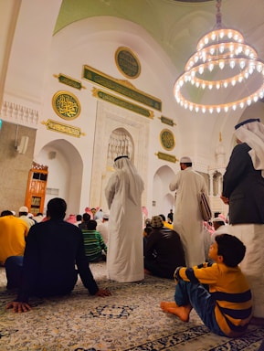 A gathering of people inside a mosque, with intricate Islamic architecture featuring Arabic calligraphy and a large chandelier hanging from the ceiling. Many of the attendees are sitting on a patterned carpet. Some are dressed in traditional Middle Eastern attire, such as white robes and keffiyehs.